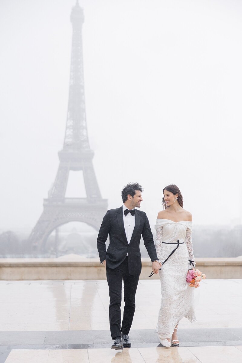 Elopement in Paris, in front of the Eiffel tower, with a bride and groom. Bride is wearing a long lace gown, the groom is in a black tuxedo