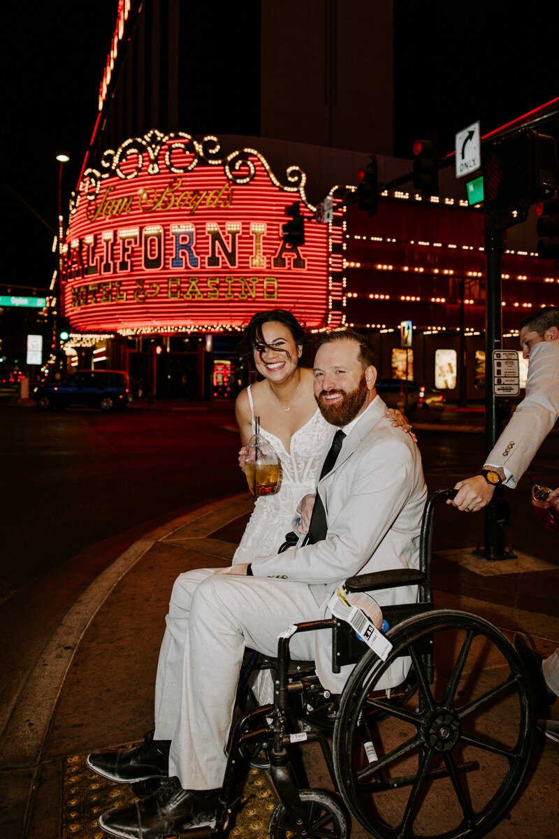 A person smiling, holding a drink, and leaning down with their arms around their partner in a wheelchair 