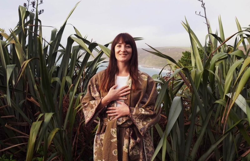 A breathing coach standing next to tall plants holding her chest and smiling