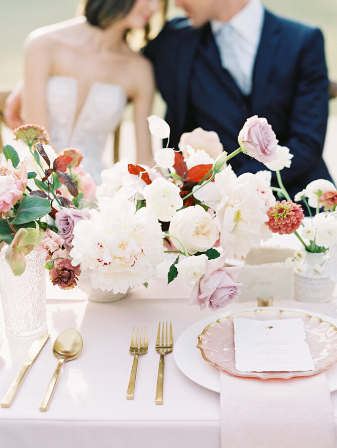 Photo of reception table setting and floral arrangement with blurry bride and groom in the background