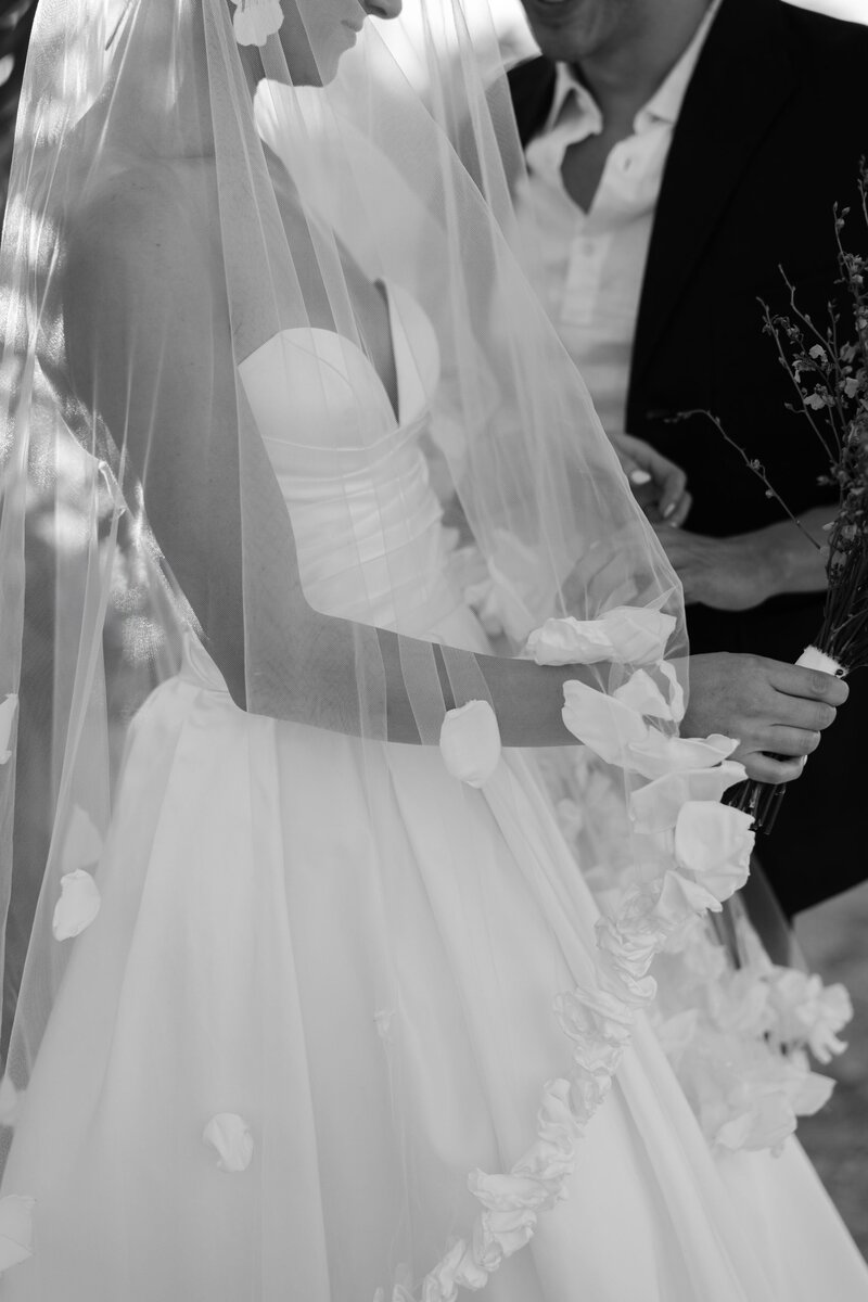 Close-up of bride and groom under soft sunlight, her veil cascading with floral appliqué.