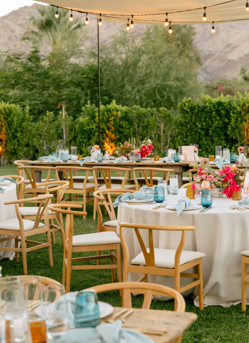 An outdoor wedding reception set up with light wooden chairs, round and rectangle tables with white table clothes, pink and red flowers, blue glasses, and a canopy above with dangling lights