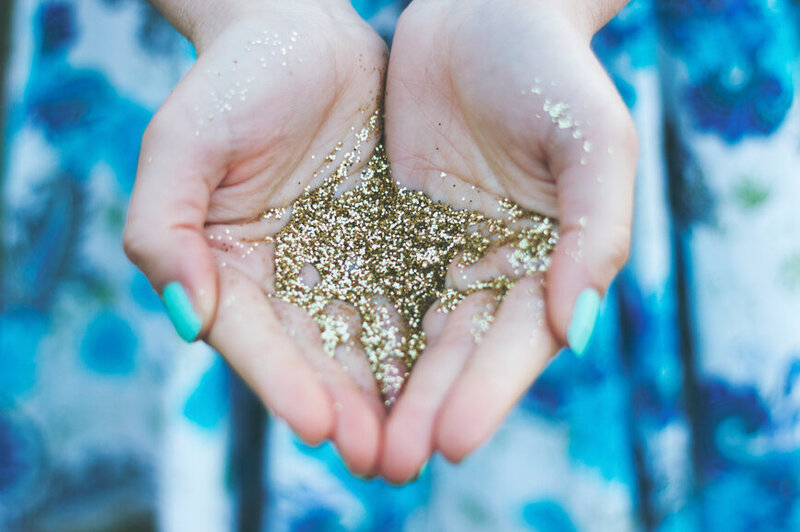 A close-up of hands holding a pile of sparkling gold glitter, with turquoise nails adding a pop of color. This image is for Debra LeClair's Embodied Style services.