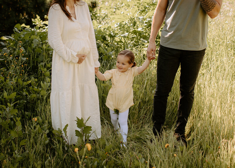 Pregnant mother holding her belly while walking hand in hand with her husband and daughter through tall grass during a spring family photo session at Horse t=Thief Canyon Park  in San Dimas, California. 