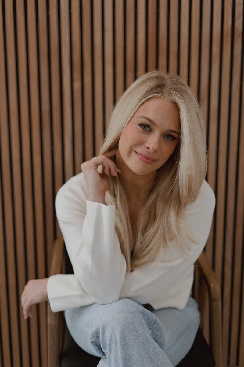 Maria Rose, founder of Sable & Rose, seated in a modern chair against a slatted wood wall, wearing a white blazer and light-wash jeans. Her long blonde hair and soft smile reflect the brand’s blend of elegance, authenticity, and approachability—symbolizing Sable & Rose’s commitment to empowering entrepreneurs with PR confidence.