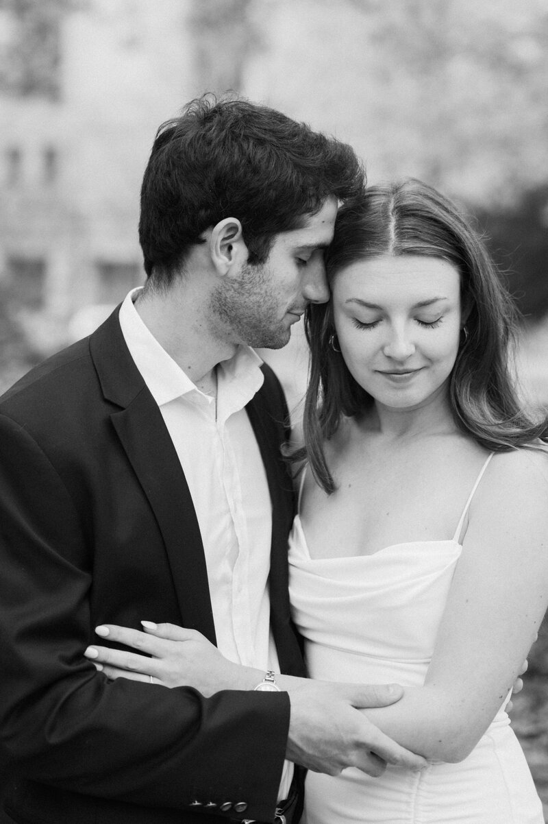 Black and white photo of a couple embracing, with the man gently resting his forehead against the woman, who is softly smiling with her eyes closed.