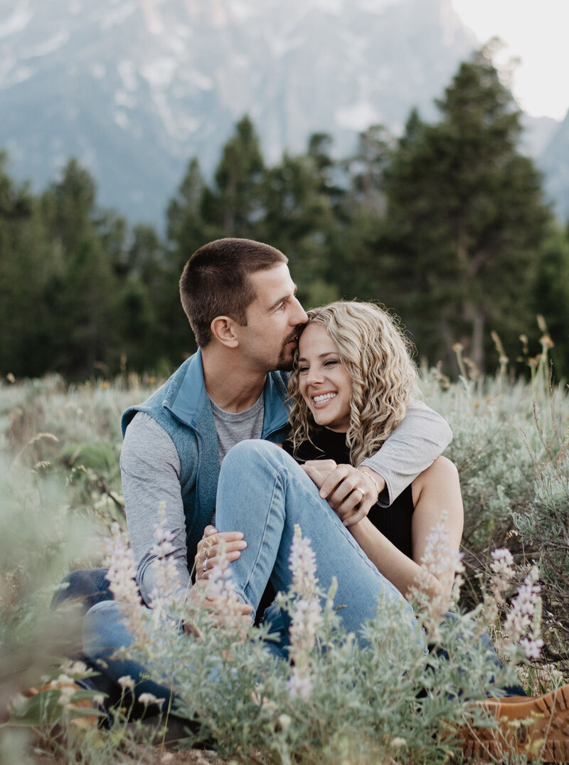 engagement photos in the grand tetons with man and woman sitting together in a field full of bushes with the mans arm wrapped around the woman and kissing her head with a lush wood behind them in the. mountains