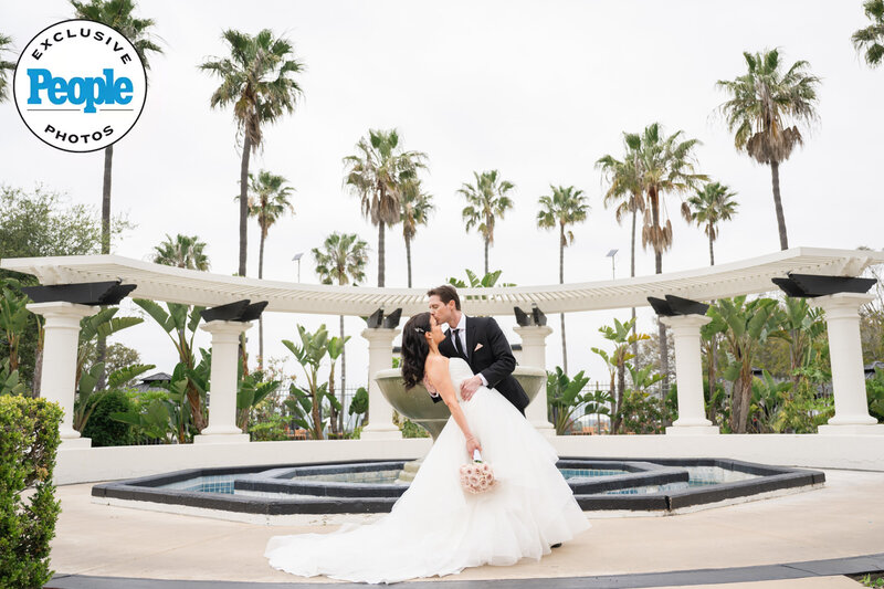 A newlywed couple shares a passionate dip kiss in front of a grand fountain at Spanish Hills, surrounded by towering palm trees and elegant architecture. The bride’s flowing gown and delicate bouquet add to the dreamy, romantic ambiance, beautifully captured by Los Angeles wedding photographers.