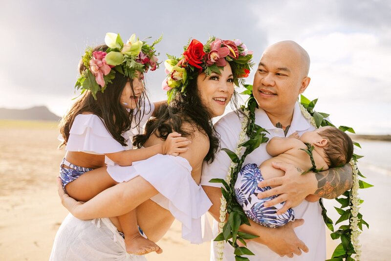 A husband and wife, with their two children wear, tropical floral, crowns and garments along a beach.