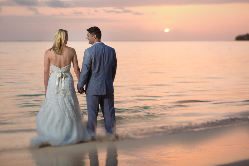 Bride and groom standing in the water at sunrise in Punta Cana, their backs to the camera as golden light surrounds them.
