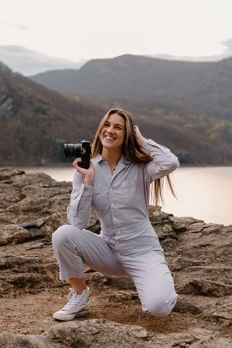 Portrait of true to color, luxury DC wedding photographer Brenna Falzone of Brenna Marie Photography posing with her camera wearing a purple long sleeve jumpsuit smiling.