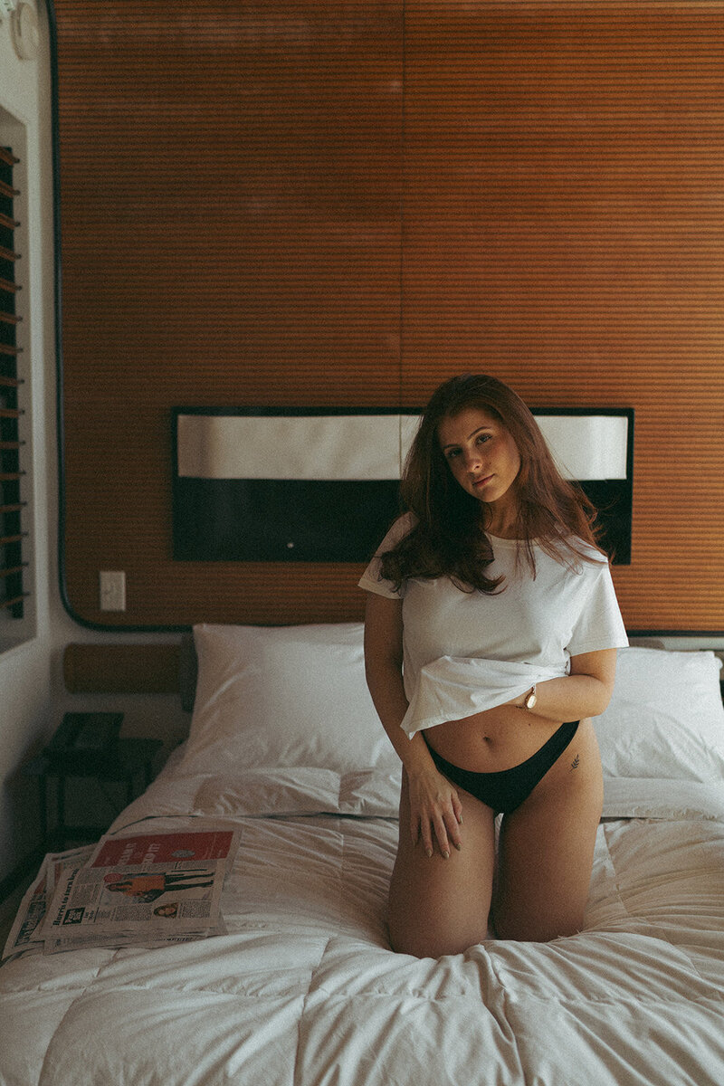 Woman kneeling on bed with hand lifting t-shirt during NYC boudoir photography session with Elsie Goodman.