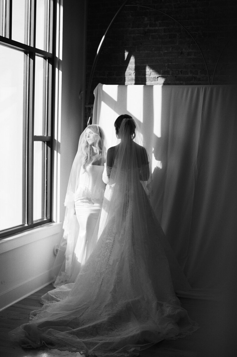 Two brides in veils standing near a window, one facing forward and one with her back to the camera.