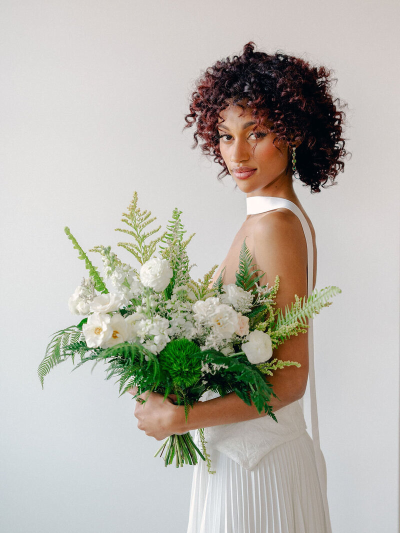 A bride poses for an intimate portrait. She is wearing a dress with a neck scarf, and holding a bouquet of elegant white and green florals.