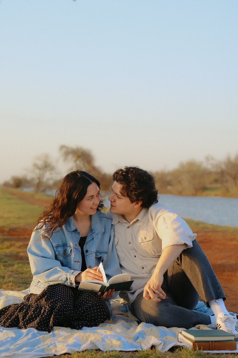 A couple looking at each other fondly as they hold a book in their hands. They are on a picnic blanket with water behind them.