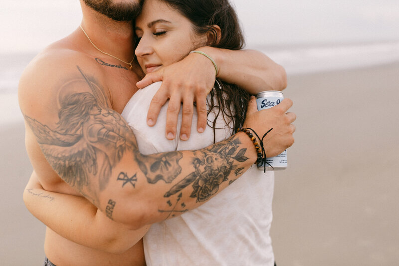 A couple hugs tightly on a beach, showcasing detailed tattoos on the man's arm against a soft sunset backdrop.