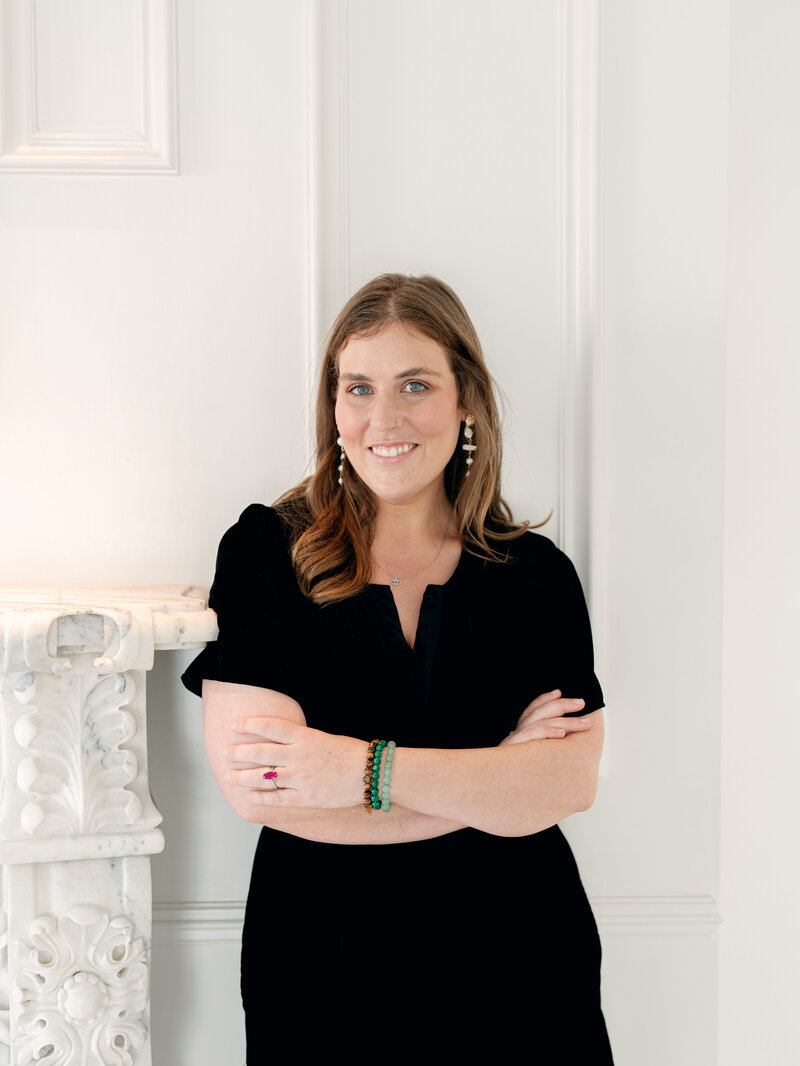 KC Cloud, smiling in a black dress standing cross armed in a white room.