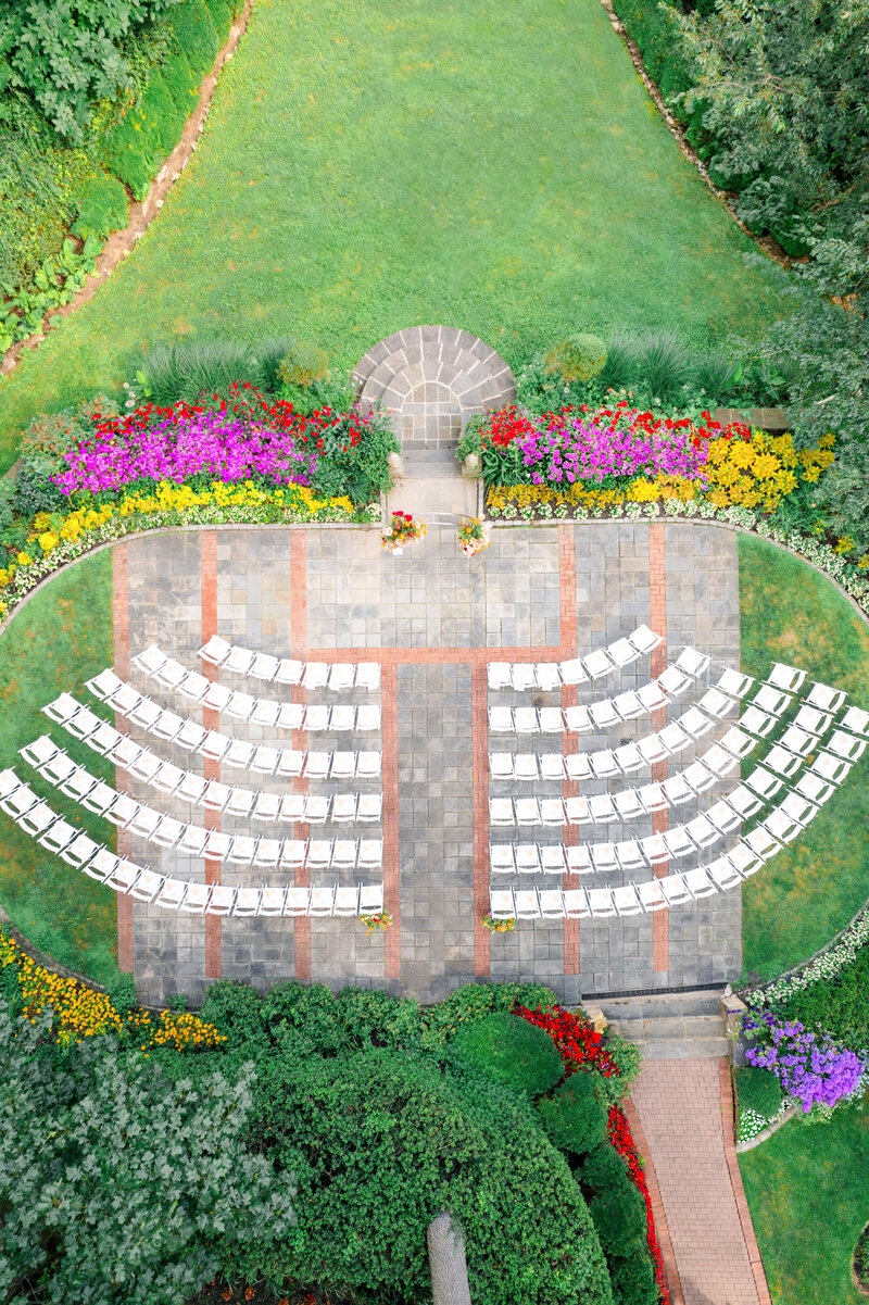 Aerial view of an outdoor wedding setup with rows of white chairs arranged in a semicircle on a stone patio. Surrounding the area are colorful flower beds and lush green grass, creating a picturesque garden atmosphere.