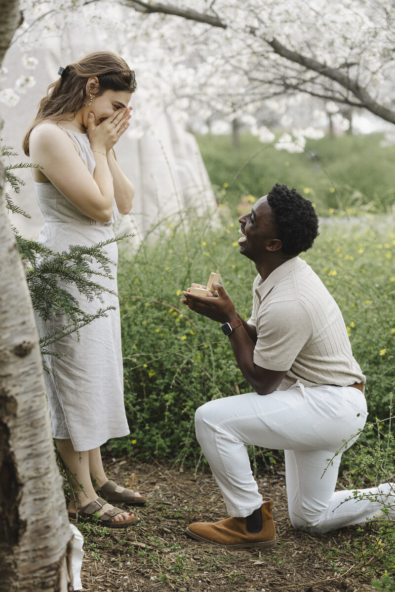 A person down on one knee proposing to their partner who has their hands over their face in surprise 