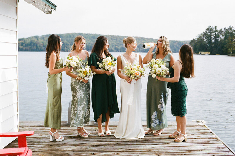 Six women in dresses stand on a lakeside dock, holding floral bouquets. The serene backdrop of calm water and tree-covered shoreline creates the perfect setting for a Toronto destination wedding photographer to capture every cherished moment.