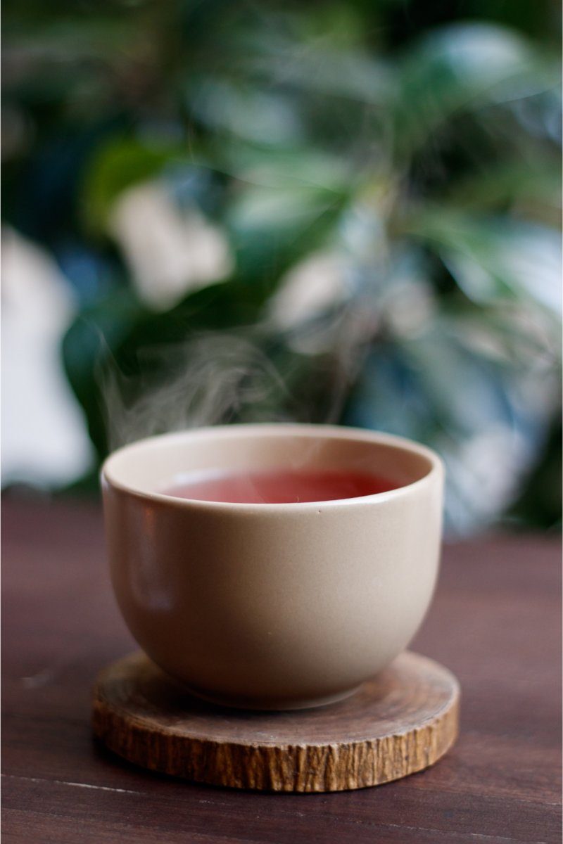white ceramic cup of hot tea on wooden coaster on brown table with blurred tree in the background