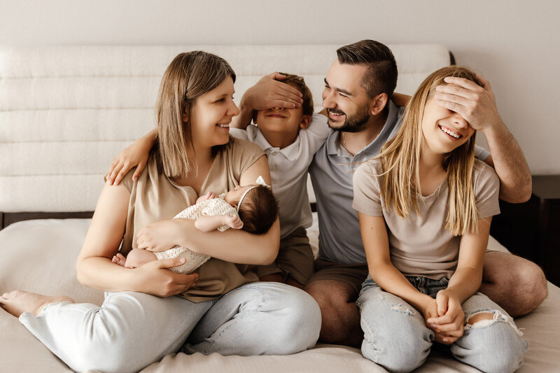 Family of five sharing a joyful moment on bed during in-home newborn photography session in Las Vegas, with parents holding their baby and playfully covering older children's eyes.