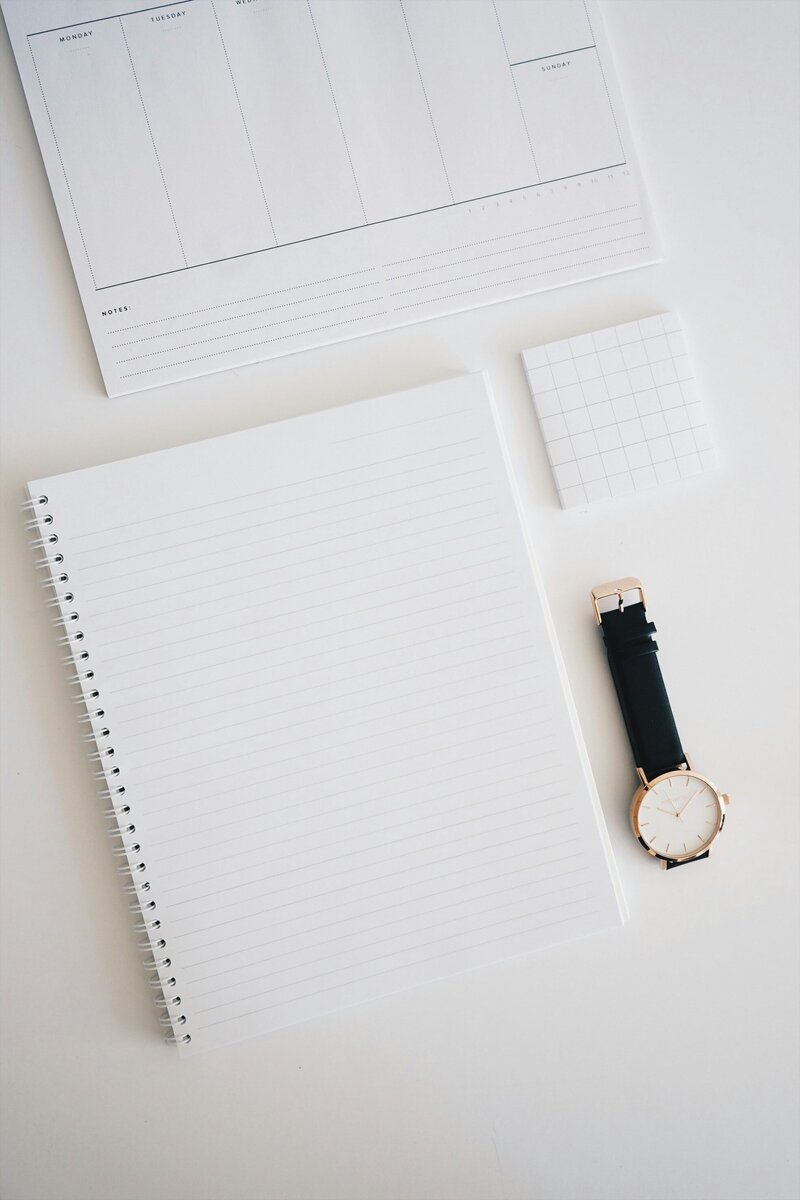 A neutral flat lay of a notebook, calendar, note pad, and watch. 