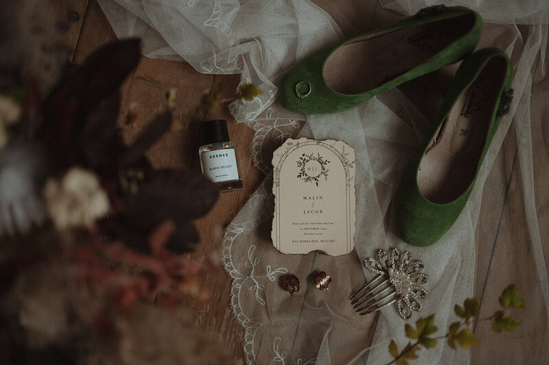 Bröllopsdetaljer utspritt på ett golv. Brudskor, vigselringar, parfymflaskor och brollopsinbjudan.