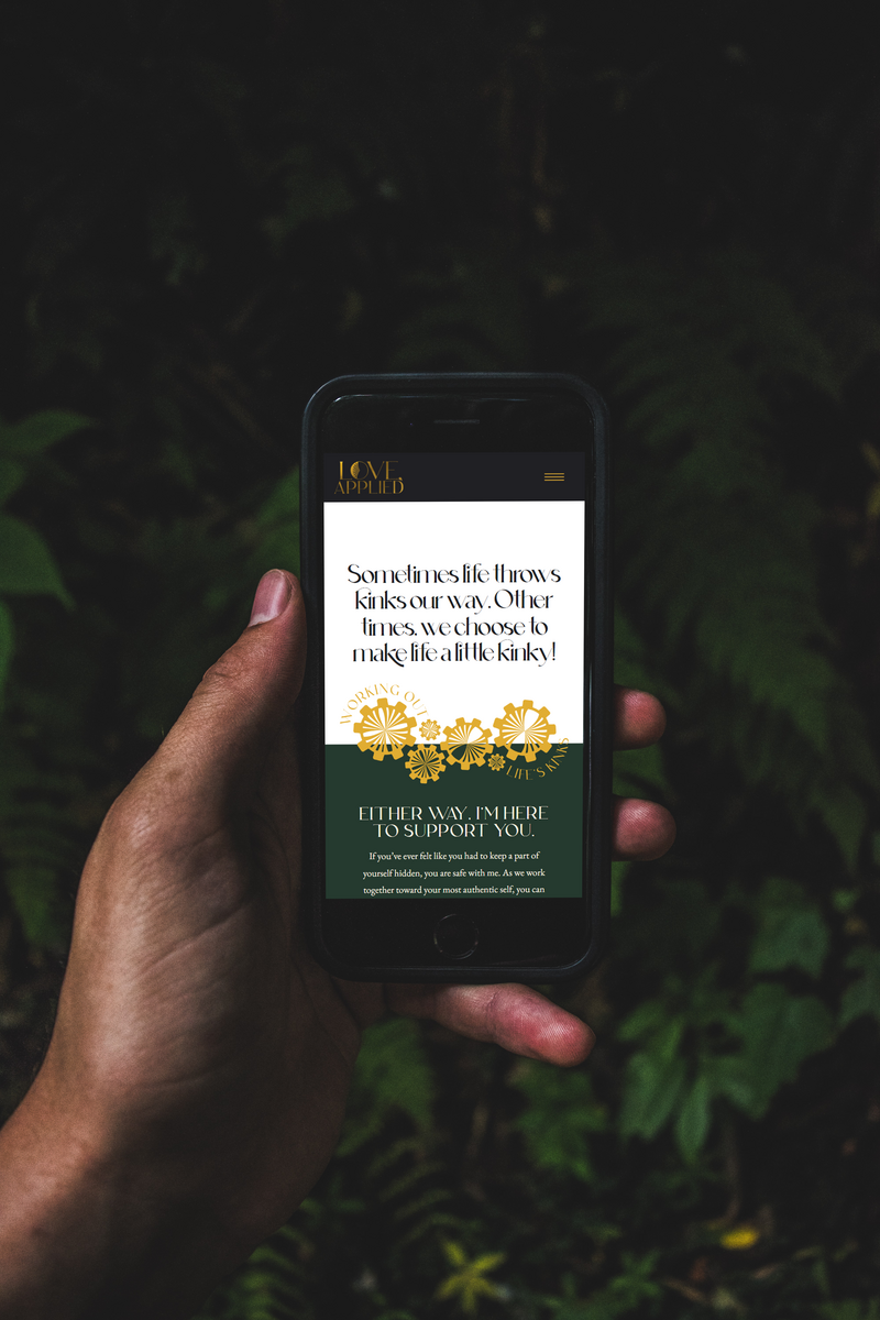 This image shows the top of the Love Applied Counseling therapy offerings page, on a smartphone with the following headline visible: "Sometimes life throws kinks our way. Other times, we choose to make life a little kinky!" Behind the phone and the hand holding it are several dark green fern leaves.