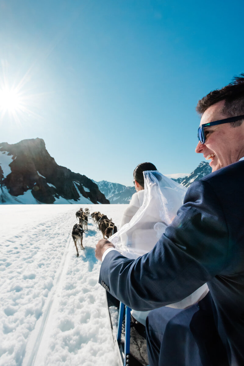 A bride and groom ride a dog sled across the glacier during their two day Kenai Fjords adventure luxe elopement.  The groom holds the bride's veil as it flies behind her in the wind.  HE is smiling broadly and laughing.  The sky is bright blue and the snow gleams white against the dark mountains rising up from the glacier.