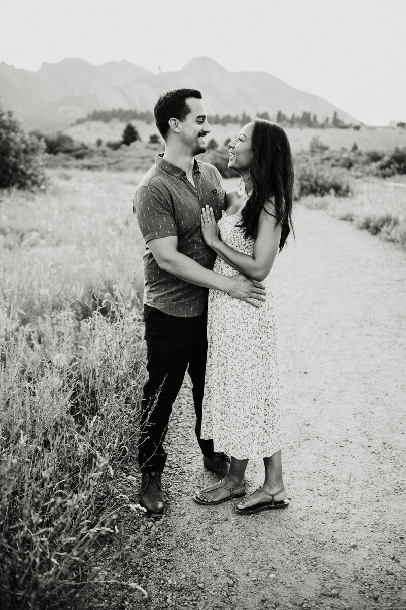 black and white shot of man wearing leather boots and button down shirt holding his fiancee as she looks lovingly at him and smiles at south mesa trail shot by colorado wedding photographer alex medvick