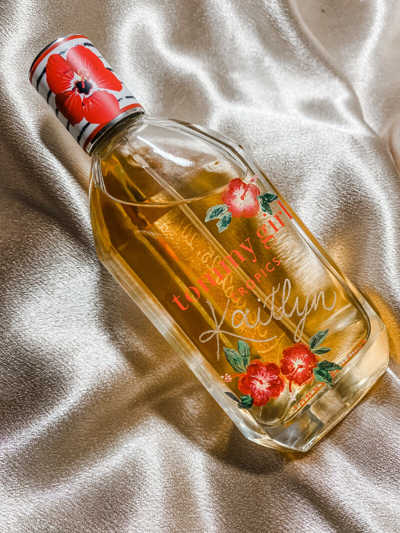 Tommy Girl perfume is lying on a chanmpagne colored silk background. It is engraved with the name "Kaitlyn" on the front with dainty red hibiscus flowers with a yellow center hand painted as a pretty embellishment. One sits in the upper right corner and two flowers on the lower left corner, below the engraved name. This was a gift for the bride from her husband.  