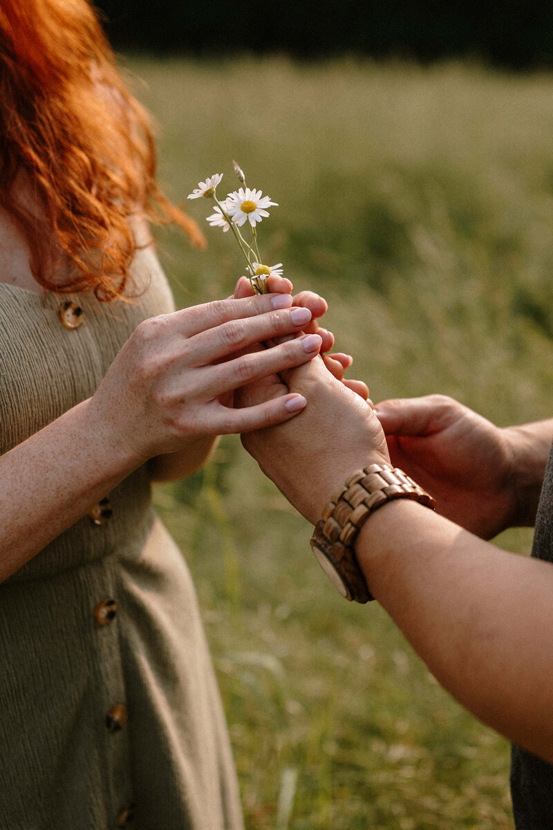 A woman with red hair gently offers a flower to a man in a grassy field, capturing a moment of connection and tenderness.