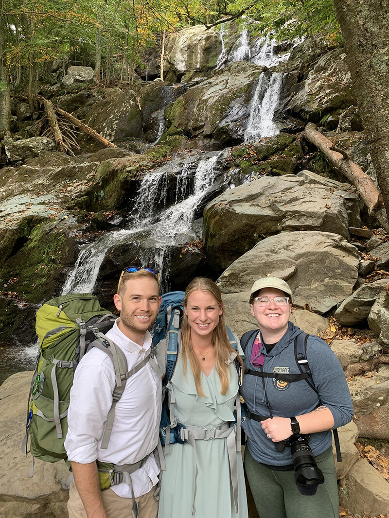 A Virginia elopement photographer stands next to an elopement couple on their wedding day. The trio stand in front of a waterfall along a trail at Shenandoah National Park.