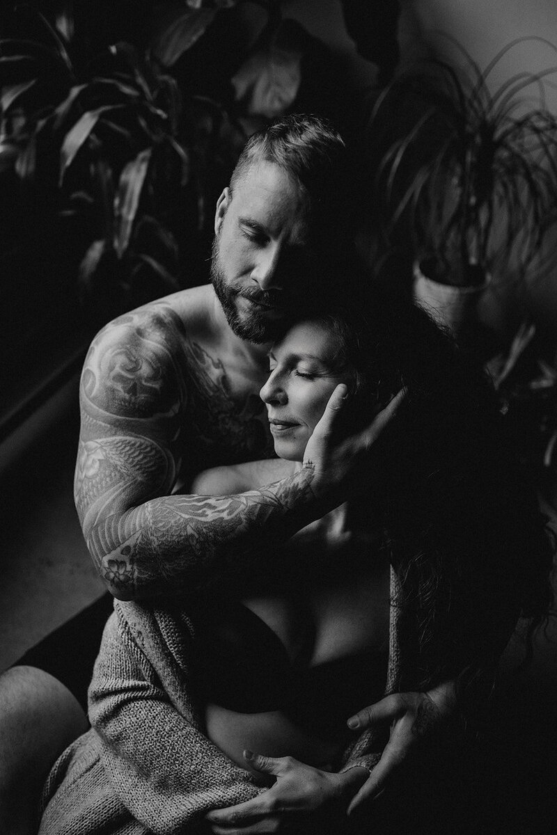 A man and a woman sit on the floor. He is behind her, his right arm reaches to the left side of her face, cupped by his hand. Her head lays on his chest with her eyes closed and head to her right. His arms and chest are covered in tattoos and he has a beard, which he rests on her forehead.