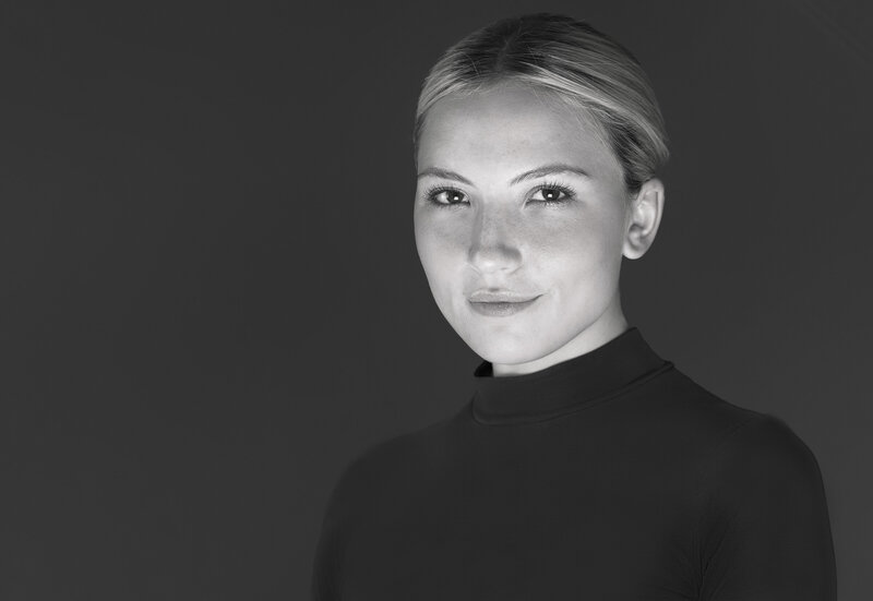 Girl in black mock neck bodysuit has her hair pulled back in a bun for a studio headshot. She is angled to the camera and has a closed-mouth smile. Philadelphia and Main Line Headshot Photographer.