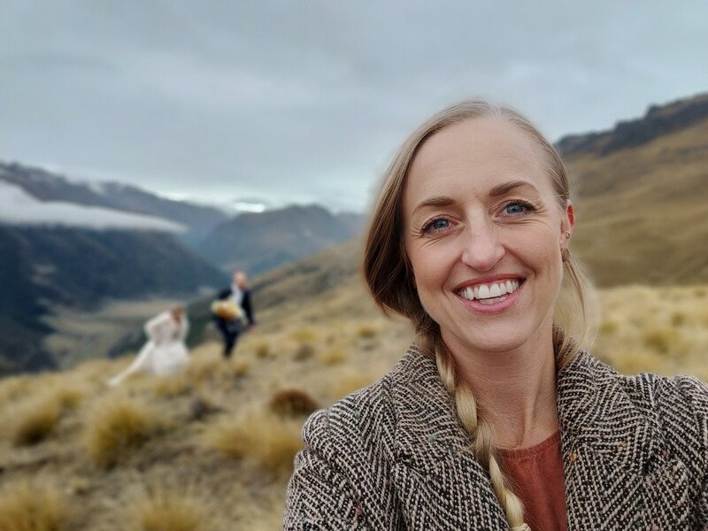 Smiling wedding celebrant taking a selfie in scenic Queenstown mountains, with bride and groom walking in the background during an adventurous outdoor elopement ceremony.