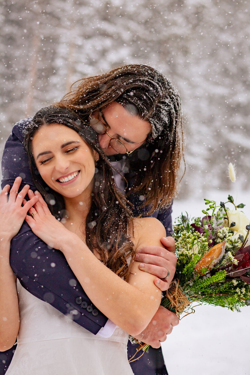 Groom hugs bride from behind at Rocky Mountain National Park during their snowy winter elopement