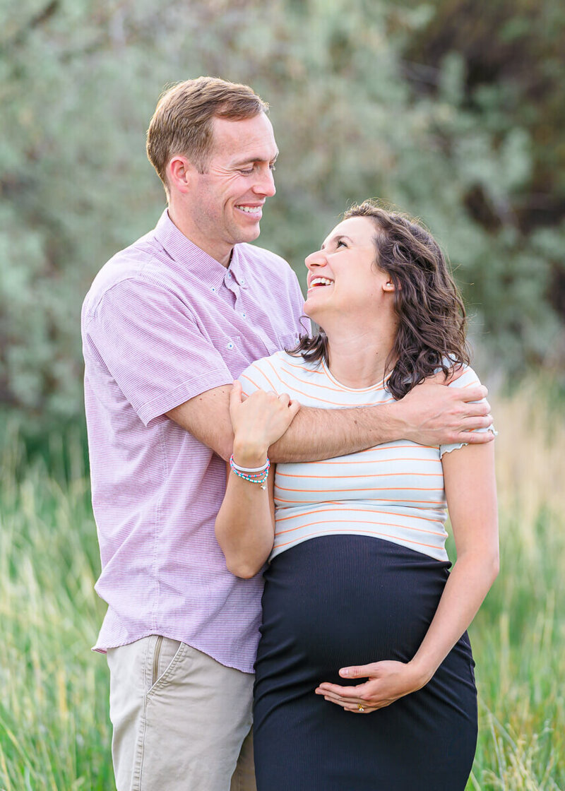 A husband wearing a pink button down shirt and khaki pants and his pregnant wife wearing a white striped shirt and black skirt smile at each other as they stand in the tall grass at North Lake Park in Lehi. Captured by Utah maternity photographer Melissa woodruff photography