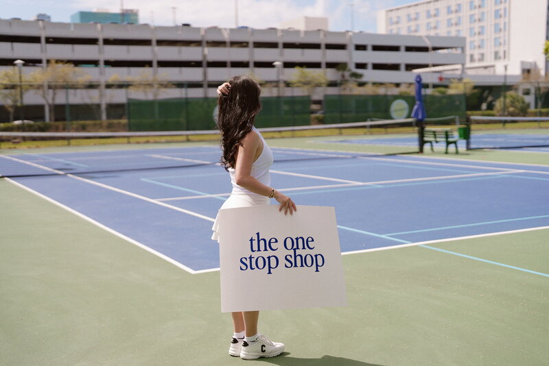 Woman on tennis court holding sign saying the one stop shop
