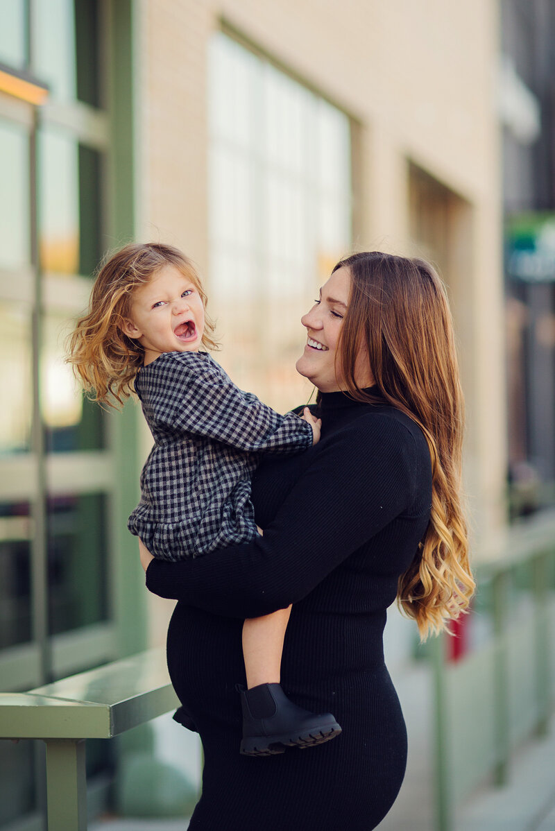 Pregnant mom is holding her toddler, who is looking at the camera smiling big. Both are wearing black near Bottleworks in Indy.