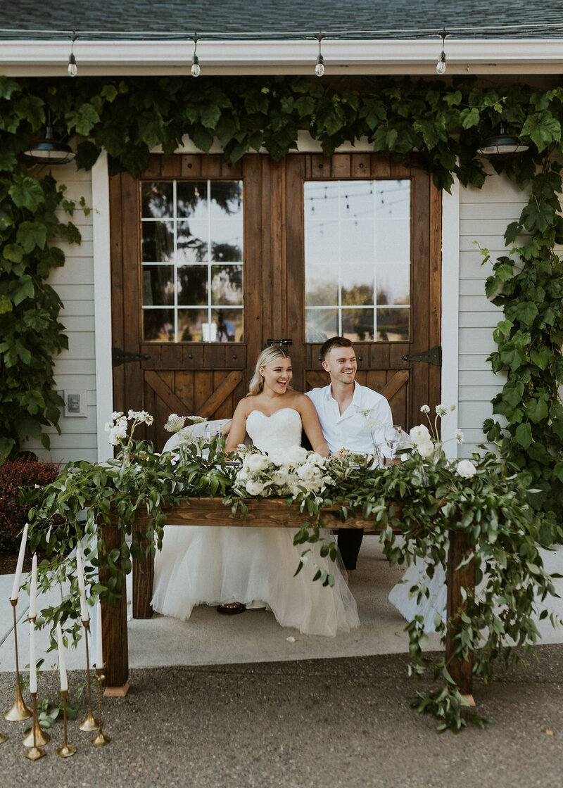 Bride and Groom sitting at wood sweat-heart table covered in greenery