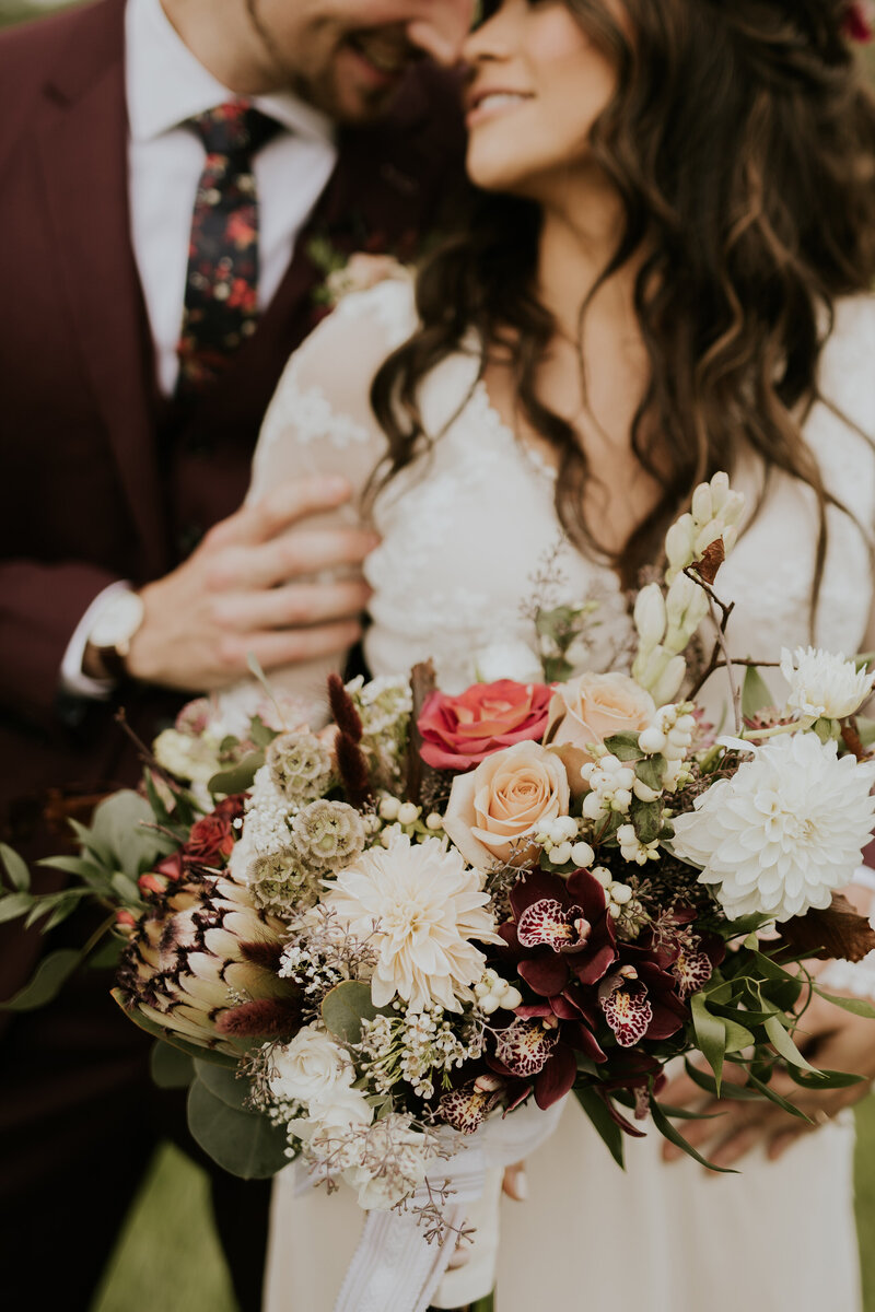 An interracial couple snuggle at their first look. The groom wears a colored suit and floral tie. The bride wears and embroidered gown and holds a bouquet of tropical flowers, dahlias, roses and orchids in rust and burgundy shades