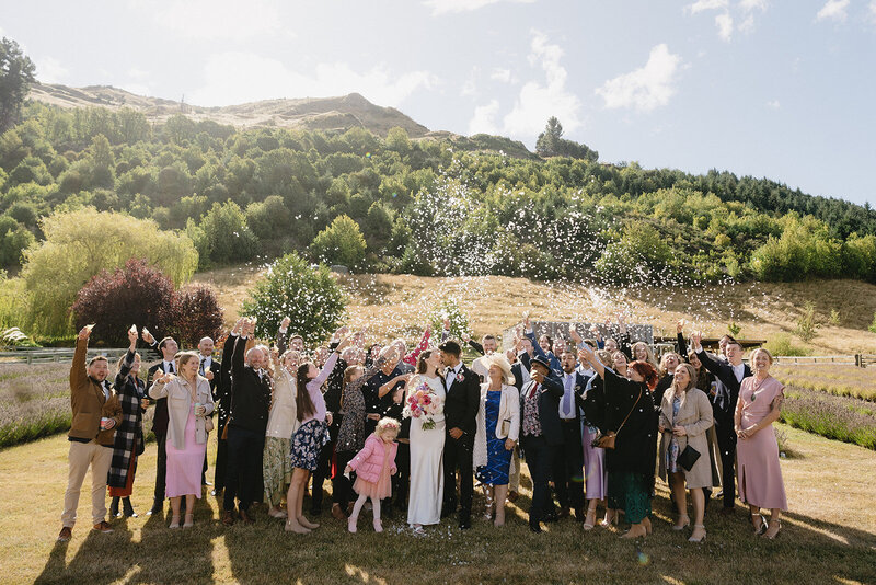 Large group of wedding guests celebrating outdoors with confetti as newlywed couple stands at the center; the background features a sunny day, rolling green hills, and a beautifully landscaped garden in Queenstown. Melissa Clark Photography