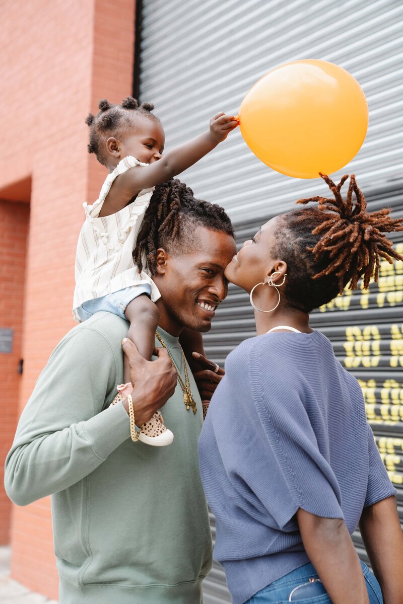 Two parents face each other, smiling, with their small child on the father's shoulders. The child holds a bright yellow balloon and smiles.