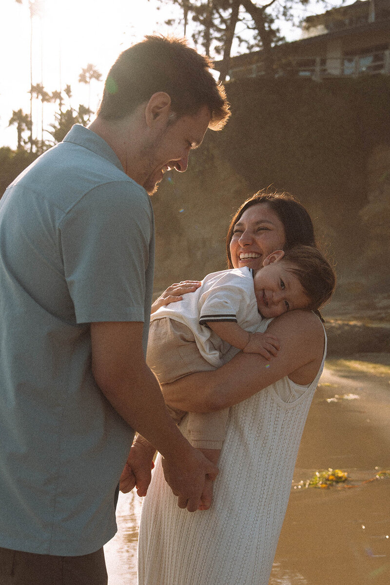 Candid family photo at Shaws Cove in Laguna Beach with parents smiling while holding their toddler during a sunset photography session.