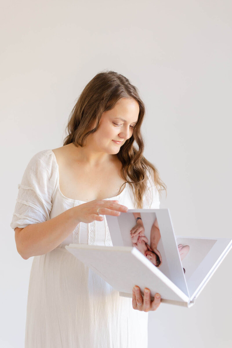 Woman in white dress with long brown wavy hair holding a newborn photo album and looking down at it as she turns the page. She is standing in an all-white photography studio.