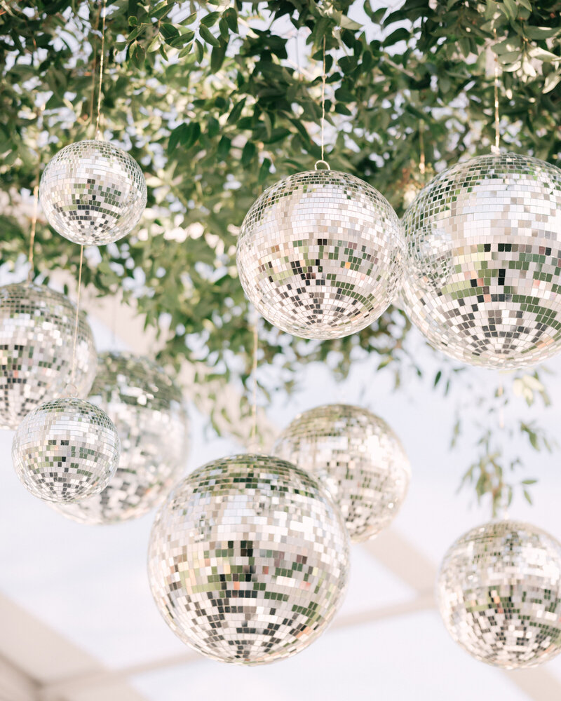 Hanging disco balls shimmer in the sunlight among leafy greenery, adding a fun and stylish touch to an outdoor wedding setting.