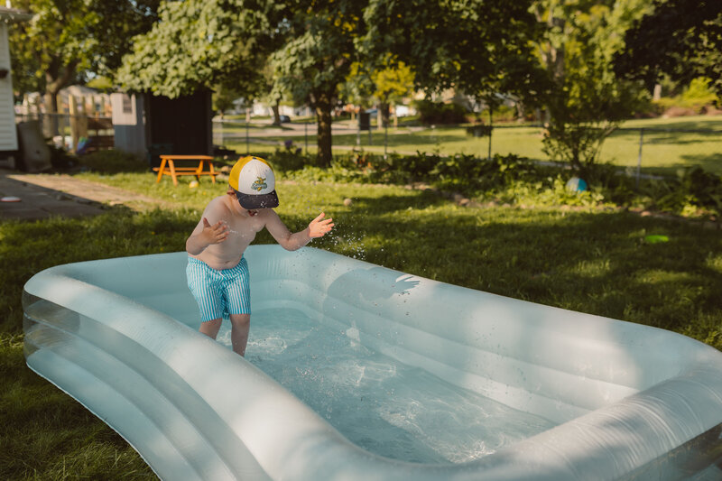 boy splashes in inflatable pool in backyard in the summer while wearing a savannah bananas hat and sunglasses