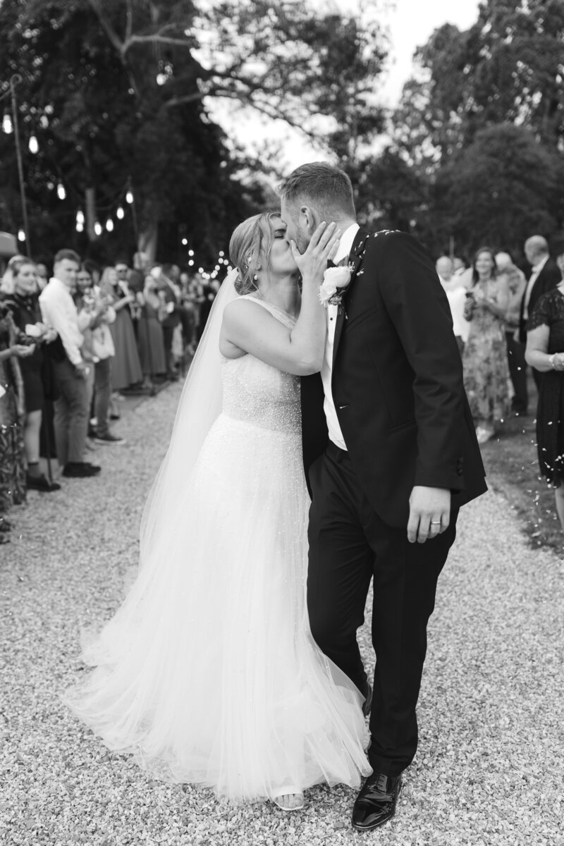 A bride in a white dress and a groom in a black suit kiss on a gravel path at Pylewell Park lined with guests.
